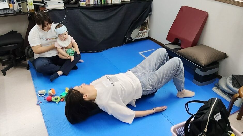 見守りスタッフがお子さんを抱っこしている横でストレッチポールでのエクササイズを行うママの様子